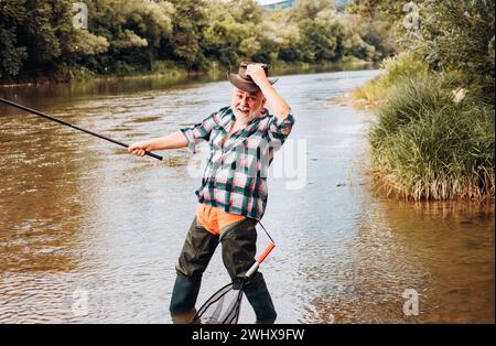 Pescatore esperto eccitato pescatore in cappello cowboy con canna da pesca, bobina rotante sul fiume. Vecchio uomo che cattura il pesce, tirando la canna mentre pesca sul lago. Foto Stock