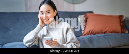 Persone e tecnologia. Donna asiatica sorridente seduta a casa, che usa il suo cellulare, digita messaggi, naviga su Internet o fa shopping Foto Stock