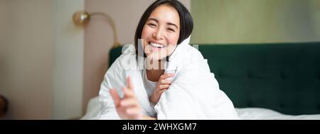 Bella donna asiatica felice coperta di coperta, siede sul letto con un piumone caldo, cerca di raggiungere la macchina fotografica con una mano, sorridendo giocherellone Foto Stock