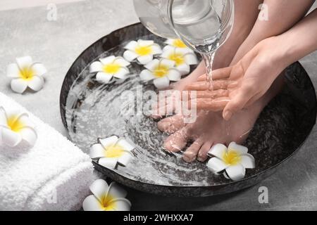 Donna che versa acqua sulla mano mentre ammorbidisce i piedi in un recipiente grigio chiaro, primo piano. Trattamento spa Foto Stock