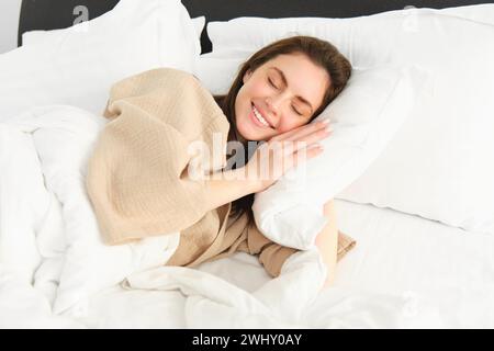 Ritratto di una bruna sorridente in pigiama, che dorme nel letto dell'hotel, che si rilassa con il viso contento, sogna, dorme in camera da letto Foto Stock