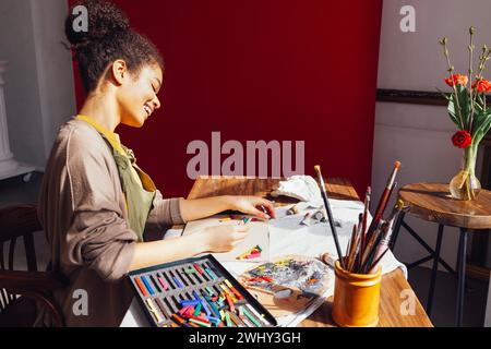 La giovane artista afro-americana, creativa e graziosa, dipinge con i colori pastello mentre si siede al tavolo davanti alla finestra Foto Stock