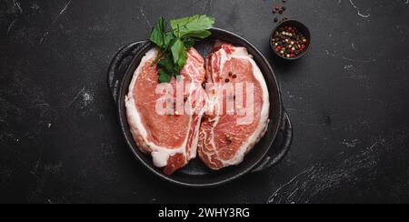 Tagliare le bistecche di maiale crude con condimenti in padella di ghisa nera su sfondo rustico scuro vista dall'alto, pronta per la tostatura. Po Foto Stock