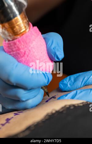 Primo piano ritagliato di una tatuatrice che lavora nel suo studio tatuando una stringa di testo sulla clavicola della sua clie femminile Foto Stock