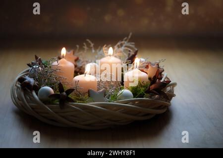 Corona dell'avvento in tonalità naturali realizzata in vimini con candele beige, muschio e decorazioni natalizie su un tavolo di legno contro il buio Foto Stock