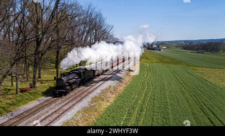 Antico treno passeggeri a vapore che viaggia attraverso le fattorie, con colture piantate, soffia di fumo Foto Stock