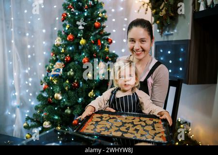 Madre sorridente con una bambina seduta al tavolo con una teglia di biscotti crudi di Natale Foto Stock