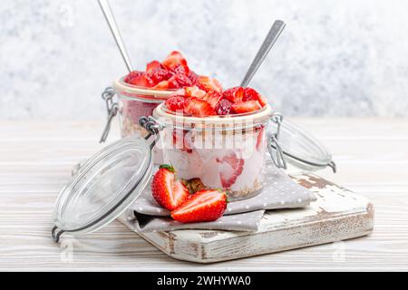 Parfait con fragole fresche, yogurt e granola croccante in vasetti da muratore in vetro trasparente su sfondo bianco rustico in legno A. Foto Stock