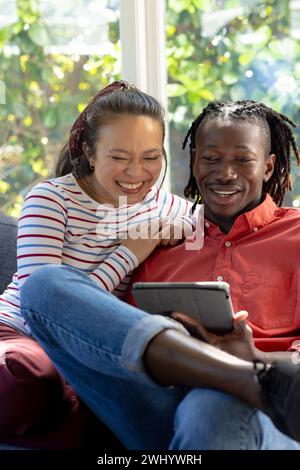 Allegri coppie diverse che usano il tablet seduto sul divano sorridendo nel soleggiato soggiorno Foto Stock