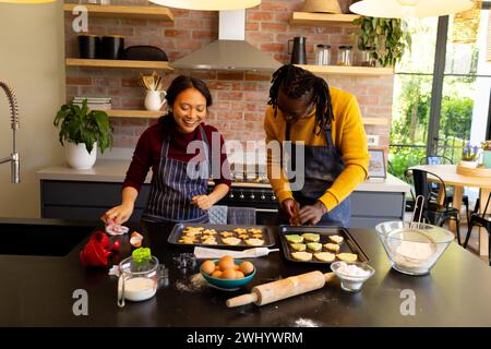 Felice coppia varia nei grembiuli che prepara biscotti di natale nella soleggiata cucina Foto Stock