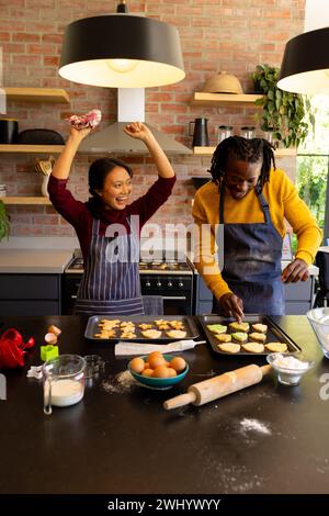 Coppia felice e varia in grembiuli che decorano biscotti natalizi e tifo in cucina Foto Stock