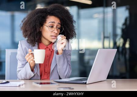 Donna malsana in posate che pulisce il viso con un tovagliolo e tiene la tazza bianca negli interni dell'ufficio. Signora ispanica che guarda lo schermo di un laptop mentre soffre di naso che cola e si prende cura con una bevanda calda. Foto Stock