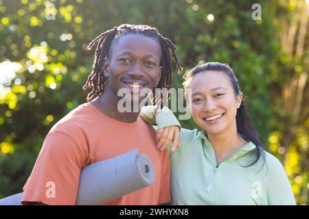 Ritratto di coppia felice e variegata che tiene in mano un tappetino yoga sorridente nel soleggiato giardino Foto Stock