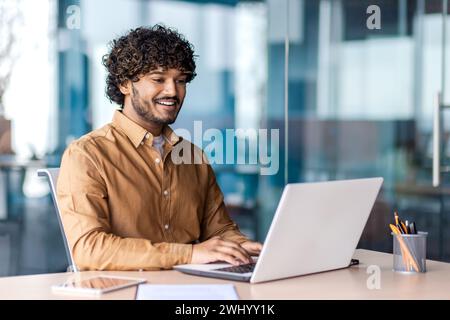 Uomo arabo di successo con la barba seduto su una sedia accanto alla scrivania con un computer portatile e un tablet accanto alle pareti di vetro. Ritratto di un giovane imprenditore che lavora online in remoto in uno spazio di lavoro condiviso per un'atmosfera produttiva. Foto Stock