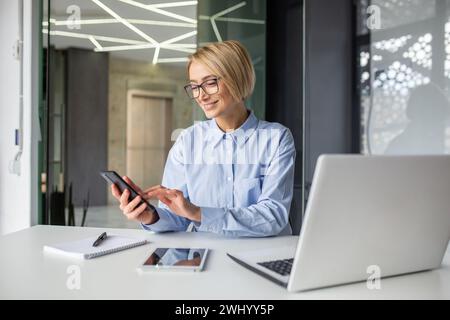Un lavoratore in ufficio che utilizza il telefono cellulare mentre si allontana dal laptop sul posto di lavoro. Donna bionda poco attenta che controlla i social media e le e-mail invece di finire il progetto aziendale. Foto Stock