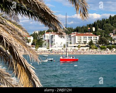 Lo yacht a vela rosso naviga sul mare sullo sfondo di una spiaggia affollata Foto Stock