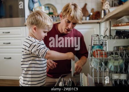 Un bambino che aiuta il padre a caricare la lavastoviglie dopo colazione. Pulire la cucina prima di andare al lavoro e all'asilo. Routine mattutina di famiglia. Foto Stock