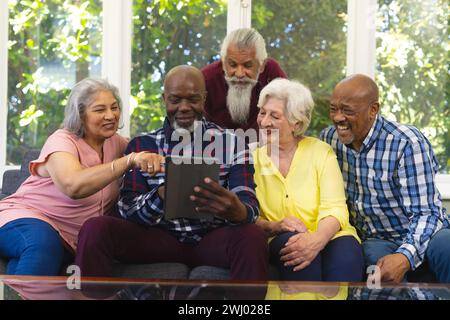 Felice e diversificato gruppo di amici anziani seduti sul divano, con tablet e sorridendo nel soleggiato soggiorno Foto Stock