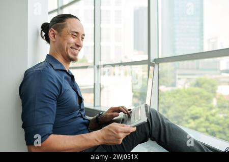 Felice giovane asiatico solopreneur maschio con laptop che digita la tastiera e guarda lo schermo mentre si siede davanti a una grande finestra Foto Stock