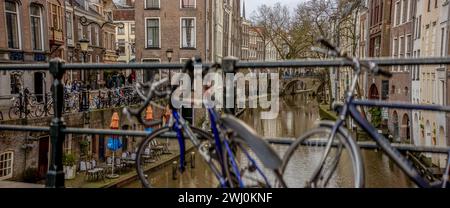 Biciclette chiuse a chiave per la ringhiera sul ponte con il canale dietro il centro storico della città olandese con negozi lungo il molo, a un livello sopra il corso d'acqua Foto Stock