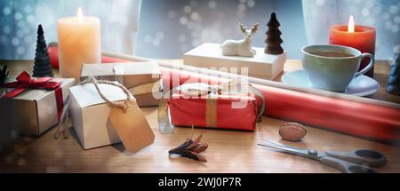 Confezione regalo natalizia su un tavolo con scatole regalo, forbici, carta rossa, candele e decorazioni natalizie, bokeh innevato, panorama Foto Stock