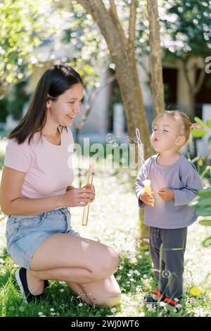 La mamma sorridente guarda una bambina che soffia bolle di sapone attraverso un bastone con molti fori Foto Stock