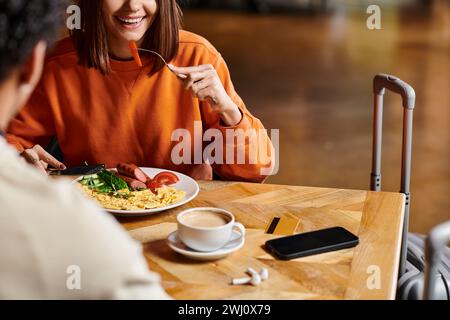 donna felice che fa colazione vicino a una tazza calda di caffè mentre si gode un pasto gustoso vicino al ragazzo nero Foto Stock