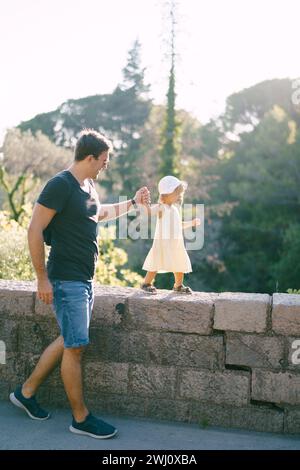 La bambina cammina lungo una recinzione di pietra nel parco tenendo il padre per mano Foto Stock