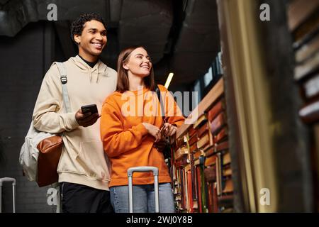 coppia felice e variegata con smartphone che condivide un momento di gioia in ostello vicino a una libreria Foto Stock