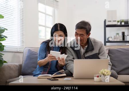 Coppie asiatiche che controllano e calcolano il conto finanziario insieme sul divano coinvolto in documenti finanziari, pagando le tasse online utilizzando il laptop a vivere Foto Stock