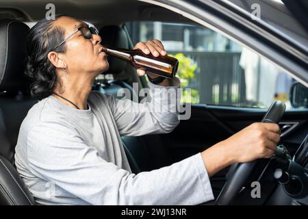 Un giovane asiatico ubriaco guida un'auto con una bottiglia di birra, un concetto di guida pericoloso. Foto Stock