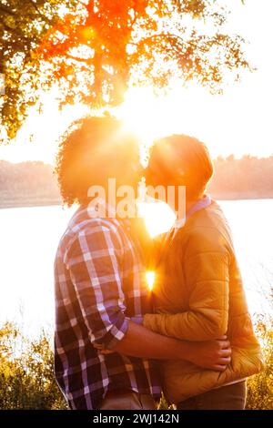 Questa immagine cattura un momento intimo tra due persone che condividono un bacio sullo sfondo di un tramonto sul lago. I raggi del sole filtrano splendidamente attraverso i rami di un albero, creando un effetto alone intorno alla coppia. Un individuo ha i capelli ricci e indossa una camicia a quadri, mentre l'altro è in una giacca gialla, e i loro profili sono illuminati dalla calda luce dorata del tramonto. La scena è evocativa di romanticismo, amore e intimità, ambientata in un ambiente naturale sereno. Romantico bacio al tramonto sul lago. Foto di alta qualità Foto Stock