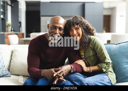 Coppia matura e diversificata che partecipa a una videochiamata, abbraccia e sorride sul divano nel soleggiato soggiorno Foto Stock
