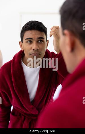 Uomo birazziale che tocca i capelli guardando nello specchio in bagno a casa Foto Stock