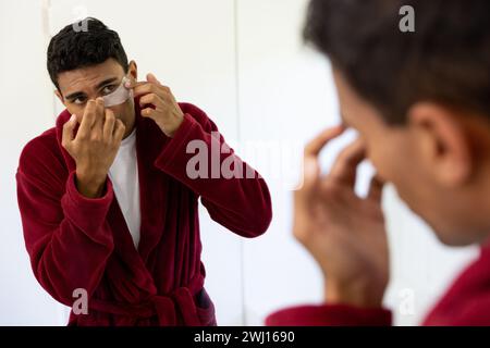 Uomo birazziale che applica cerotti sotto gli occhi che guardano nello specchio in bagno a casa Foto Stock