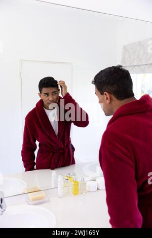 Uomo birazziale che tocca i capelli guardando nello specchio in bagno a casa Foto Stock