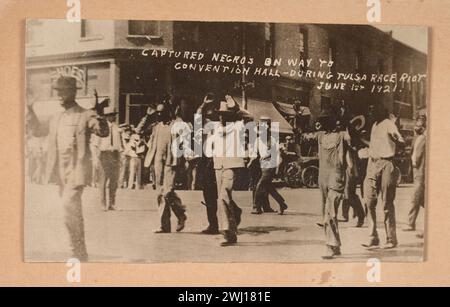 TULSA RACE RIOT (31 maggio e 1 giugno 1921): Una fotografia in bianco e nero di uomini che camminano in mezzo a una strada con le mani alzate in resa durante il massacro di Tulsa nel 1921. Sullo sfondo, una folla di persone fiancheggia la strada di fronte a un edificio. Nel negativo della fotografia è scritto [CATTURATO NEGROS SULLA STRADA PER / SALA CONVEGNI - DURANTE LA RIVOLTA DI TULSA / 1 GIUGNO 1921.]. Foto Stock