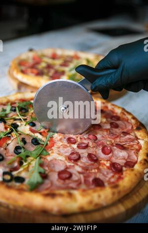 Tagliare la pizza a fette con un coltello speciale. Pizza fresca sullo sfondo. Coltello con le mani. Foto Stock