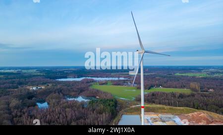 Questa immagine aerea presenta una turbina eolica solitaria che domina il primo piano, con le sue pale che raggiungono un cielo spazioso. Sotto, un mosaico di foreste e campi si estende in lontananza, intervallato da piccoli corpi d'acqua che riflettono il cielo nuvoloso. La turbina è un simbolo della moderna energia rinnovabile, in contrasto con il paesaggio naturale sottostante. La fotografia cattura l'intersezione tra l'innovazione umana e la bellezza della natura Vista aerea della turbina eolica solitaria che si affaccia sulla foresta. Foto di alta qualità Foto Stock
