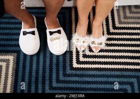 I piedi maschili e femminili nelle ciabattine si trovano su un tappeto a righe. Didascalia: Sposo. Sposa. Tagliato, senza volto Foto Stock