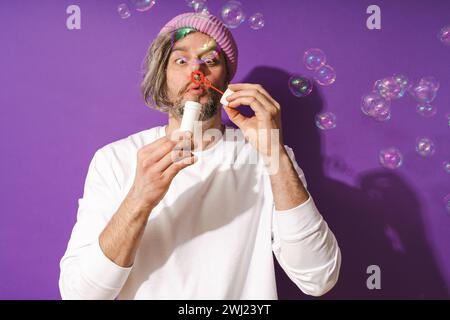 Uomo di mezza età spensierato che soffia bolle di sapone su sfondo viola Foto Stock