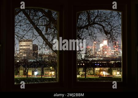 Viste attraverso una finestra a bovindo di una casa residenziale e i rami degli alberi sono le finestre illuminate delle case d'epoca terrazzate che confinano con il parco Ruskin di Lambeth, e in lontananza una nuova torre a Higgs Yard a Loughborough Junction (a sinistra) e lo sviluppo a Nine Elms (a destra), l'11 febbraio 2024, a Londra, Inghilterra. Foto Stock