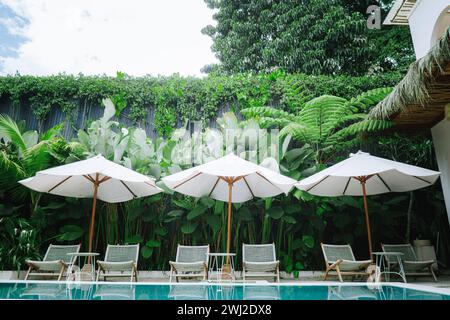 Lussuosa area piscina tropicale all'aperto circondata da vegetazione lussureggiante e da un cielo azzurro limpido con tre ombrelli bianchi con pali di legno aperti. Foto Stock