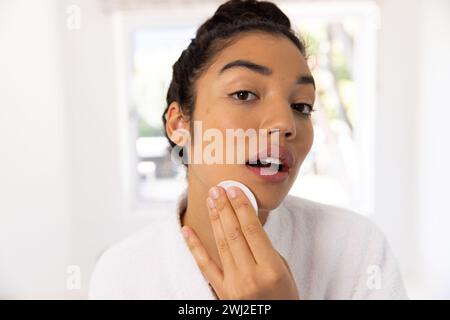 Ritratto di donna birazziale in accappatoio che pulisce il viso in un bagno soleggiato Foto Stock