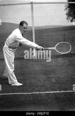 Anni '1920, storico, all'aperto su un campo da tennis in erba, un tennista gentiluomo con l'elegante abito sportivo dell'epoca - pantaloni lunghi in flanella, maglietta a maniche lunghe e cravatta - che gioca a dorso con una tradizionale racchetta in legno, Inghilterra, Regno Unito. Foto Stock