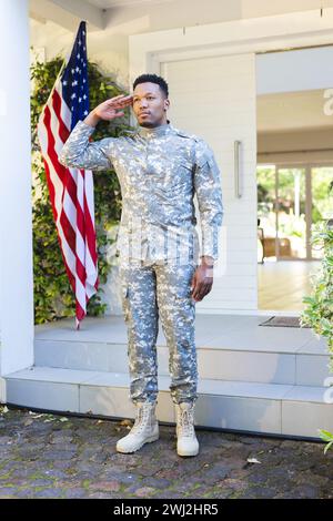 Soldato afroamericano concentrato in piedi e salutando fuori casa con la bandiera degli stati uniti, spazio copia Foto Stock