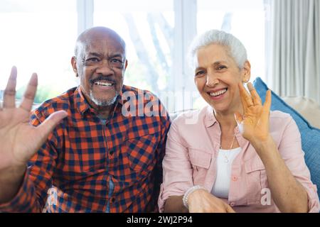 Coppia senior felice e variegata che effettua videochiamate in un soggiorno soleggiato Foto Stock