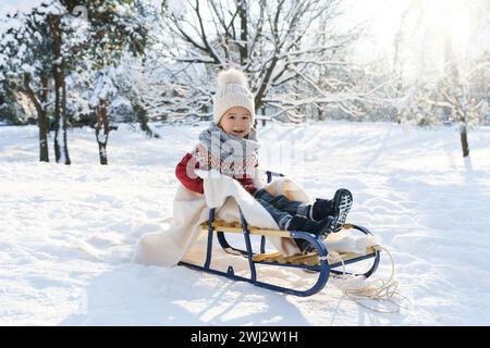 Bambino seduto sulla slitta in un parco cittadino innevato durante le soleggiate giornate invernali Foto Stock