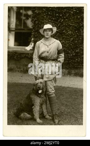 Cartolina originale dell'era WW1 di una ragazza di casa chiamata Esme Greene che indossa una tunica beige che denota i doveri generali della fattoria (al contrario dell'uniforme bianca per il volo) e una fascia da braccio di servizio di 3 mesi. Indossa i breeches, una tunica totale al ginocchio (con cintura). Si trova fuori da una casa di campagna o fattoria, con un cane chiamato Kim, datato 1917, Easton a Gordano, vicino a Bristol, Regno Unito Foto Stock