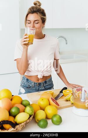 Donna che beve succo d'arancia appena spremuto in casa in cucina bianca Foto Stock